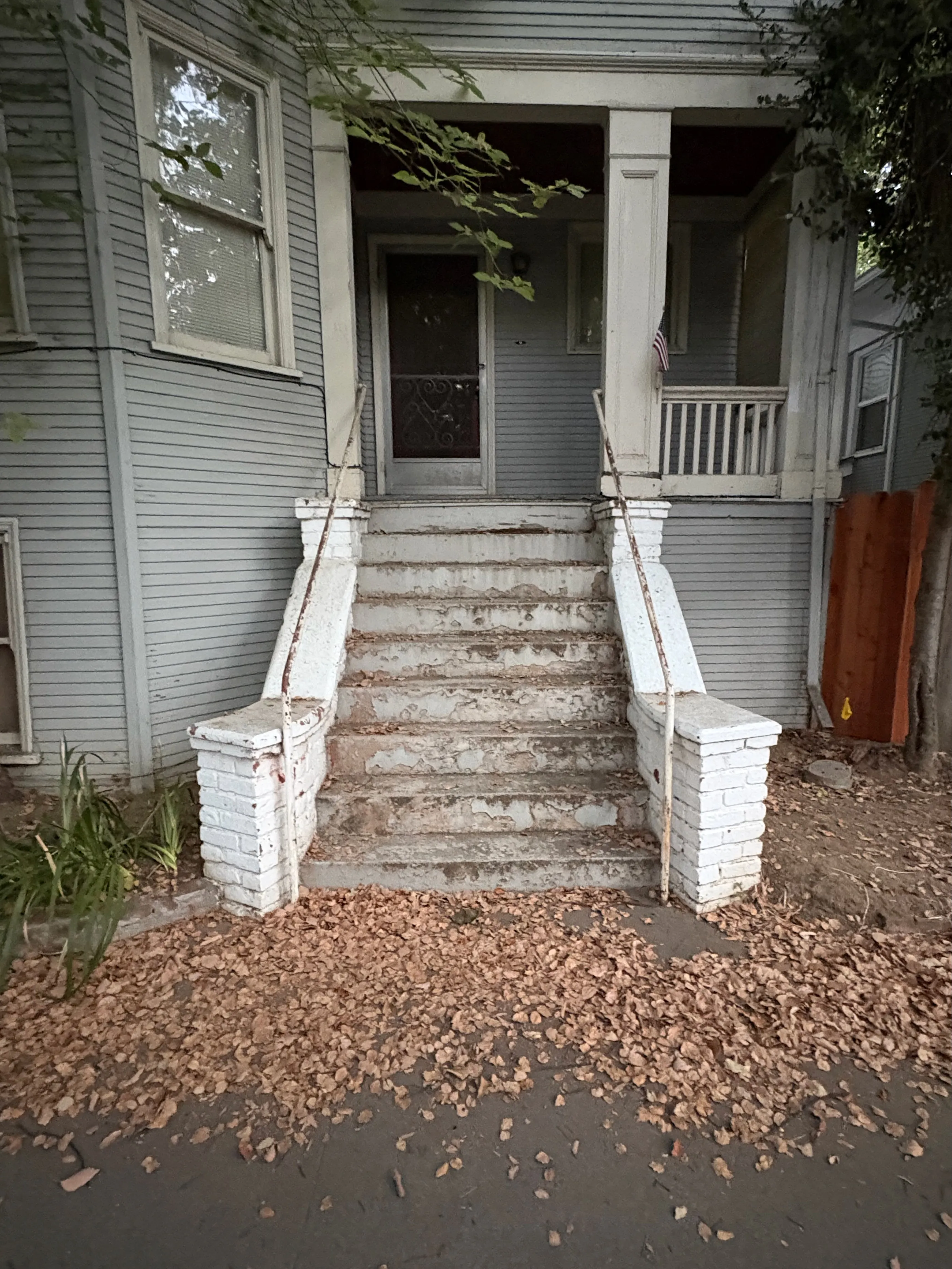 Brown, fallen leaves are strewn at the base of an aging brick and cement set of stairs in the foreground. Nine stairs in total lead up to the small, covered front porch of a similarly aging apartment building. The wood siding slats are a powdery, barely blue hue. The shadowy porch is unoccupied, save for a small American flag that protrudes from a support pillar in the direction of the camera.